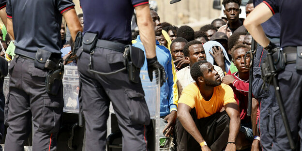 Italienische Carabinieri stehen vor Migranten, die auf dem Boden sitzen