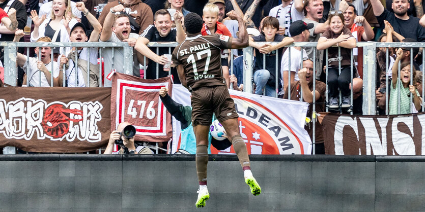 ein Fußballspieler in braunem Dress springt vor Fans in die Luft und reckt die Faust