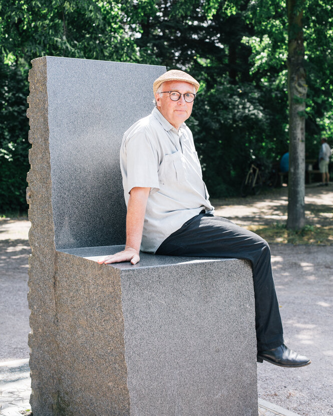 Daniel Gaede sitzt auf dem Goethe-Hafis-Denkmal. Das Denkmal ist aus Stein und ähnelt einem überdimensionalen Stuhl. Gaede trägt ein helles Hemd, eine schwarze Hose, einen hellen Hut und eine Brille.