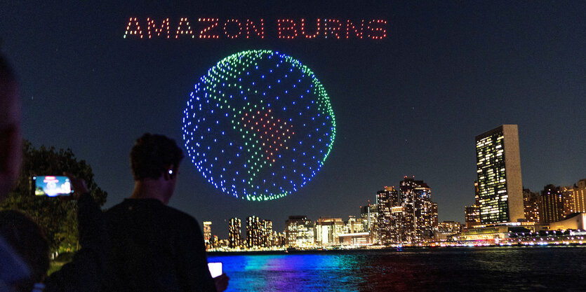 Menschen beobachten Leuchtbild der Erde am Himmel mit Schriftzug "amazon burns"