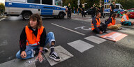 Aktivistin mit Warnweste sitzt auf der Straße mit weiteren Aktivistinnen und hat sich festgeklebt , im Hintergrund ein Polizeiwagen und mehrere Polizisten
