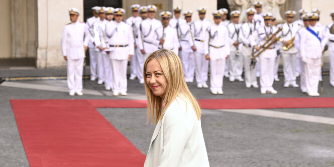 Premierministerin Giorgia Meloni, mit blonden Haaren und einer weißen Jacke, vor einer Kapelle.