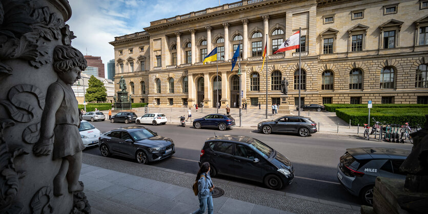 Das Abgeordnetenhaus wird von der gegenüberliegenden Straßenseite gezeigt. Vor dem Haus laufen Menschen und auf der Straße stehen Autos.