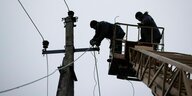 Zwei Arbeiter reparieren in luftiger Höhe in einem Kranwagen stehend eine zerstörte Leitung an einem Strommast