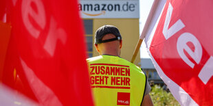 Im Hintergrund eine Amazon Betriebsstätte, im Vordergrund ein Mensch in Ver.di-Warnweste, mit Ver.di-Fahne