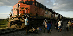 Ein großer Güterzug wird seitlich von der Sonne beschienen, vor ihm stehen mehrere Menschen mit großen Rucksacken
