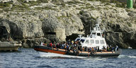 Gerettete Migranten sitzen auf einem Boot der italienischen Küstenwache auf dem Weg zum Hafen von Lampedusa. Das Boot fährt an der felsigen Küste vorbei