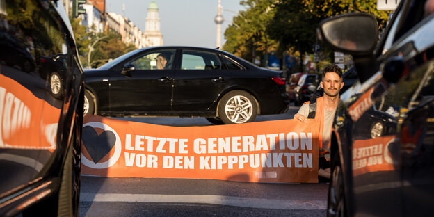 Ein Aktivist der Letzten Generation sitzt mit einem Transparent der Klimabewegung in der Hand auf der Straße vor dem Brandenburger Tor und behindert den Straßenverkehr. Vor und hinter ihm stehen Autos.