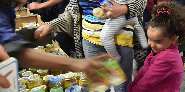 Ein kleines Mädchen steht an einem Tisch mit Babygläschen, eine Mutter hält ihr Baby auf dem Arm
