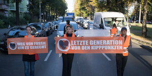 Drei Aktivist*innen der Letzten Generation laufen mit einem Banner vor Autos her. Auf den Bannern steht: "Letzte Generation vor den Kipppunkten" und "Weg von Fossil, hin zu gerecht".