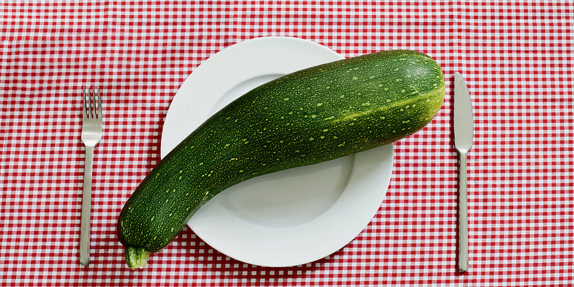 Eine große Zucchini liegt auf einem Teller. Besteck liegt daneben