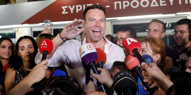 Stefanos Kasselakis steht lachend mit geöffnetem Hemdkragen vor Mikrofonen der Presse