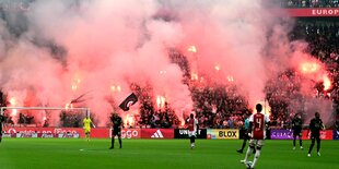 Fußballspieler auf dem Rasen, im Hintergrund Bengalos auf den Zuschauerrängen