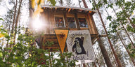 Protestcamp im Wald, mit einem Baumhaus hoch in den Bäumen, an dem ein Protestplakat hängt: Wuhlheide bleibt