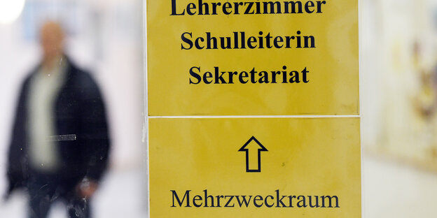 Ein gelbes Schild an einer Glastür zeigt den Weg zur Schulleitung. Dahinter steht ein Mann.