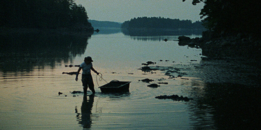 Eine Fru im Morgenlicht mit den Füßen im See, an der Hand eine Art Wasserschlitten