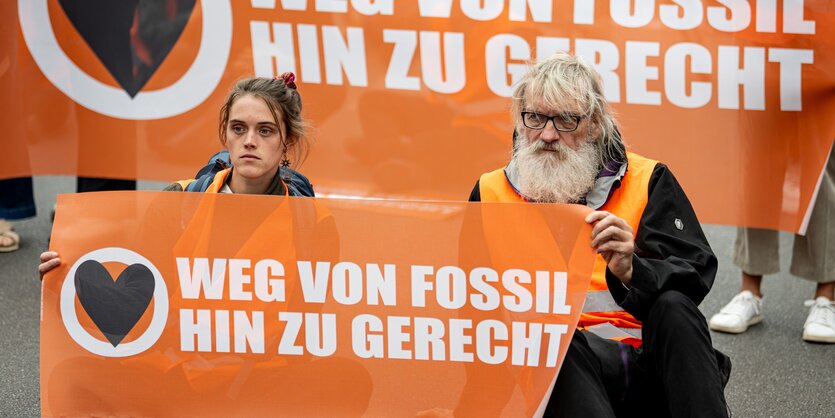 Zwei Aktivist*innen der Letzten Generation sitzen, ein Banner haltend, nebeneinander auf der Straße.