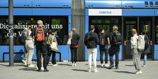 Studierende, fast alle haben Rucksäcke auf, stehen vor einer einfahrenden Tram