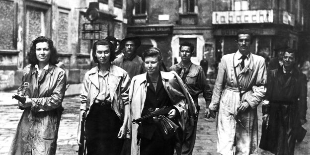 schwarz weiss Foto aus den 40er Jahren. Drei Frauen und ein Mann tragen helle Mäntel und gehen mit Waffen die Straße entlang