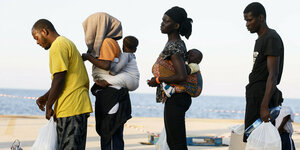 Vier Migranten warten mit Gepäck und Kindern auf dem Rücken an einer Mole auf Lampedusa