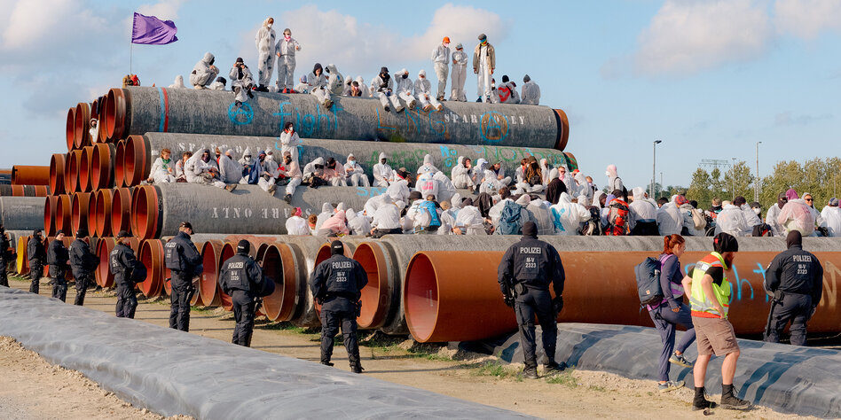 Menschen in weißer Schutzkleidung sitzen auf großen Rohren, vor ihnen stehen Polizisten