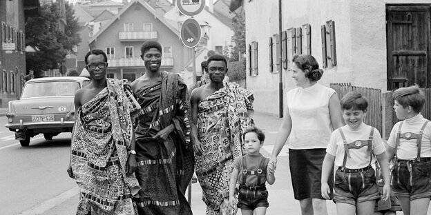 Drei Männer aus Ghana mit traditionellen Gewändern gehen mit einer Frau und ihren drei Kindern, die Lederhosen tragen, durch Murnau in den 60iger Jahren
