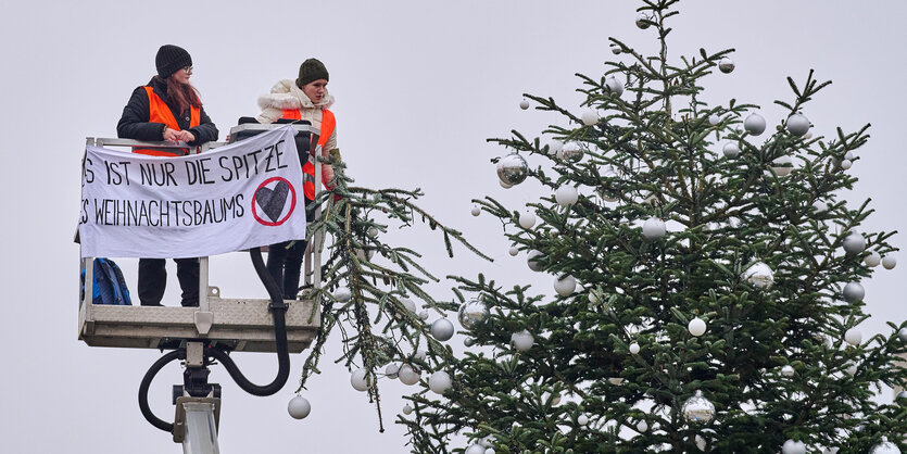 Zwei Aktivistinnen der Letzten Generation stehen mit Warnwesten auf einer Hebebühne hoch oben neben dem Weihnachtsbaum: ein Transparent ist an der Hebebühne befestigt: Es ist nur die Spitze des Wihnachtsbaums