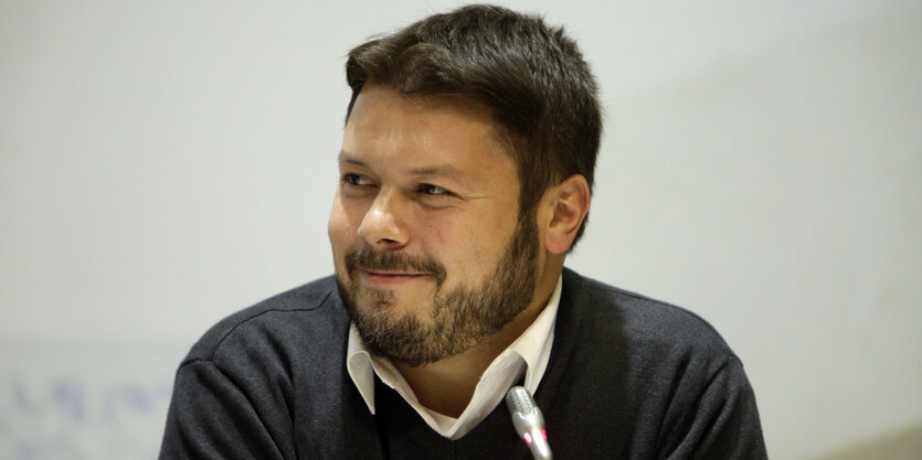 Ender Cetin lächelt auf einer Pressekonferenz, vor ihm steht ein Mikro auf dem Tisch.