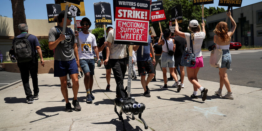 Ein Roboterhund trägt ein Protestschild undd läuft mit einer Gruppe von menschen über einen gehweg