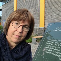 eine Frau mit Brille neben einer Tafel, die auf den Gedenkort für deportierte Flensburger Sinti und Roma hinweist