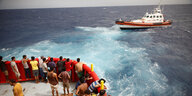 Geflüchtete stehen an der Reling eines Rettungsschiffs, ein zweites kleineres Boot ist ebenfalls auf dem Meer zu sehen