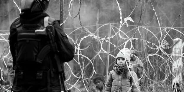 Ein Kind in Winterkleidung steht hinter einem Stacheldrahtzaun, vor dem ein bewaffneter Soldat steht.