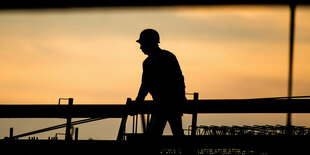 Die Silhouette eines Bauarbeiters zeichnet sich auf einer Baustelle vor dem verfärbten Morgenhimmel ab.