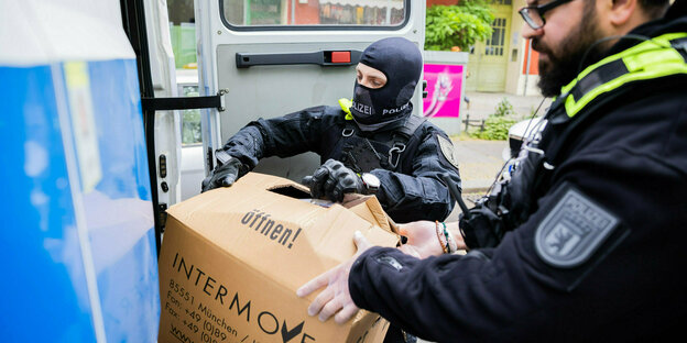 Zwei Beamte wuchten einen Karton in ein Fahrzeug, einer trägt eine Maske über dem Gesicht