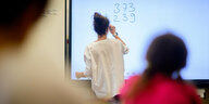 Schülerin von hinten, sie blickt auf ein Smartboard, eine Lehrerin schreibt dort Zahlen an