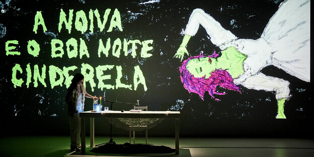 In Neonfarben ist eine schlafende Frau dazu der Titel der Performance auf portugiesisch auf eine schwarze Wand pojiziert. Davor eine Frau, die sich gerade auf einem Tisch stehende Getränke einschenkt