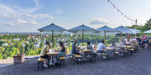 Menschen sitzen in einem Gartenlokal mit Blick über einen Hügel und Wiesbaden