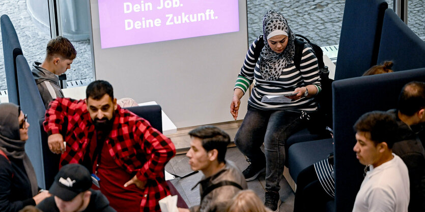 Menschen sitzen in einer Halle und informieren sich über Jobs. Im Hintergrund ist ein Schild mit der Aufschrift Dein Job, deine Zukunft