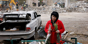 Eine Frau steht trauernd mit Blumen in der Hand am Rand einer Straße, die von Zerstörung gezeichnet sit. Im Hintergrund ein resttlos zerbombtes Gebäude, die dem Hilfstrupps stehen