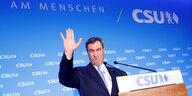 Markus Söder hebt die Hand vor einem Rednerpult. Er sieht etwas zerknautscht aus. Hinter ihm eine blaue Wand mit dem CSU Logo und der Behauptung: Am Menschen