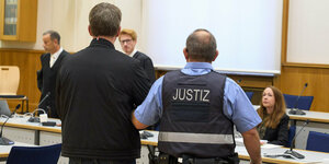 Zwei Männer sind von hinten zu sehen, der eine trägt eine schwarze Jacke, der andere ein Hemd mit der Aufschrift "Justiz"