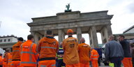 Eine Gruppe Entsorgungskräfte in orangenen Jacken steht mit dem Rücken zur Kamera vor dem Brandenburger Tor