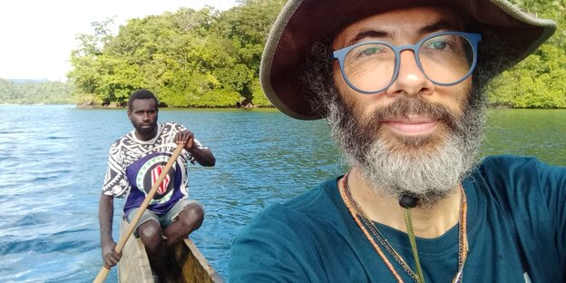 Selfie: weißer Mann mit Hut, Bart und Brille, aufgenommen in einem schmalen Holzkanu, im Hintergrund ist ein Mann zu sehen, der das Ruder eintaucht