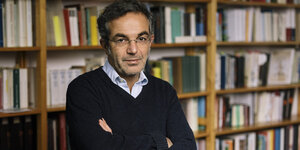 Navid Kermani steht in einem dunklen Pulli vor einer Bücherwand und verschränkt die Arme