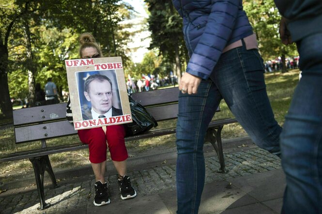 Eine Frau sitzt mit einem Plakat auf einer Bank, Menschen gehen vorbei. Das Plakat zeigt das Porträt von Donald Tusk mit der Inschrift: Ich vertraue dir, Donald