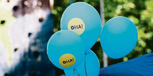 Vier hellblaue Ballons mit Aufschrift "Oha" schweben in der Luft.