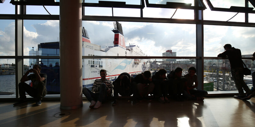 Flüchtlinge sitzen auf dem Fußboden einer Wartehalle. Im Hintergrund ist die Fähre zu sehen