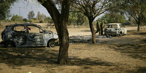 Zwei ausgebrannte Autos auf einem Gelände mit Bäumen und zwei Soldaten. Die Soldaten schauen in ein Auto