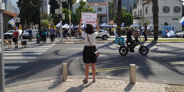 Eine Frau steht auf einer Straße in tel Aviv und hält ein Schild in die Höhe, darauf steht geschrieben "Bring back the hostages"