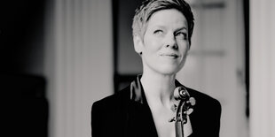 Schwarz-Weiß-Porträt von Isabelle Faust. Die Violinistin trägt ein schwarzes Jacket und kurze Haare. Sie schaut nach oben. Vor ihr ragt der obere Hals einer Violine ins Bild.